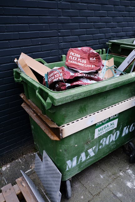 A large green wheeled bin positioned outdoors on a paved surface next to a dark brick wall, filled with packaging materials including cardboard boxes and red plastic bags containing construction or garden supplies labeled 'FK200 FLEXY PLUS.' Nearby, a white plastic crate with green markings supports the bin, which appears to be part of home relocation or waste disposal services. The items inside the bin suggest materials collected during packing or renovation activities, with some materials protruding from the top. The surrounding area includes a metal ramp or platform on the ground, indicative of a loading process, potentially managed by a professional removals service like Man with Van Northolt to facilitate efficient furniture transport and overall moving logistics.