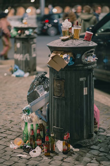 A public street scene showing a large, black plastic waste bin with a dome-shaped lid, situated on cobbled pavement. The bin is overflowing with discarded paper, cardboard, plastic bottles, and food packaging, with some rubbish spilling onto the ground. Several empty glass bottles, including beer and soft drink bottles, are scattered around the base of the bin. On top of the bin, there are crumpled paper cups, a food container, and a paper bag, indicating recent waste disposal. In the background, blurred pedestrians and parked cars are visible, suggesting an active urban environment. The scene highlights typical street litter near roadside parking, relevant in the context of removal and disposal services like those offered by Man with Van Northolt to assist with home relocation or bulk waste clearance, ensuring proper disposal to avoid fines.