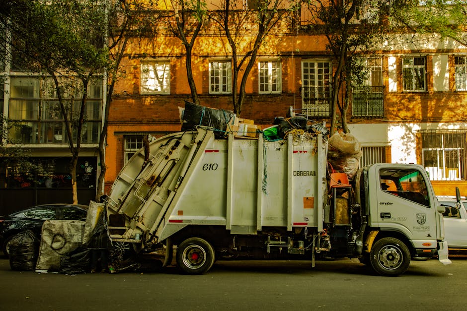 A large green wheeled bin positioned outdoors on a paved surface next to a dark brick wall, filled with packaging materials including cardboard boxes and red plastic bags containing construction or garden supplies labeled 'FK200 FLEXY PLUS.' Nearby, a white plastic crate with green markings supports the bin, which appears to be part of home relocation or waste disposal services. The items inside the bin suggest materials collected during packing or renovation activities, with some materials protruding from the top. The surrounding area includes a metal ramp or platform on the ground, indicative of a loading process, potentially managed by a professional removals service like Man with Van Northolt to facilitate efficient furniture transport and overall moving logistics.