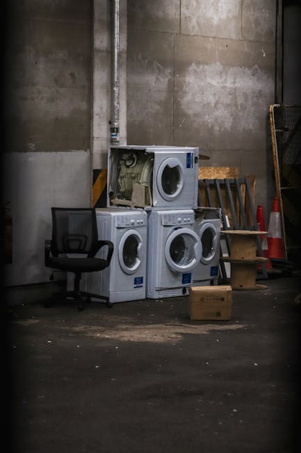 In an industrial or storage area with concrete walls and a concrete floor, four white domestic washing machines are stacked together, with two placed side by side on the floor and two positioned on top of each other. The door of the topmost washing machine is open, revealing the drum inside. Nearby, there is a black office chair with a mesh backrest, and a wooden step ladder leaning against the wall behind the appliances. To the right of the stacked washing machines, red and white traffic cones are placed, and a small cardboard box is positioned on the floor in front of the appliances. The scene appears to be part of a home relocation or furniture transport process, with the washing machines prepared for loading onto a van by Man with Van Northolt, a professional removals company specializing in house moves and packing logistics.