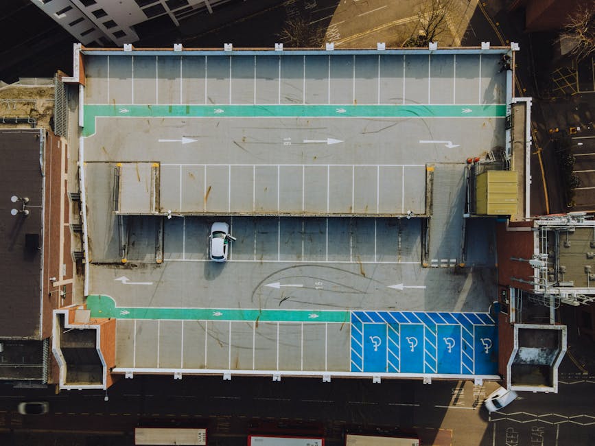 An aerial view of a rooftop parking area demonstrating a designated loading zone with white painted arrows indicating vehicle movement directions. The parking area contains multiple clearly marked spaces, including blue zones reserved for disabled parking, and a section with green markings. A small white van is parked in the middle of the lot, surrounded by various materials such as wooden pallets, cardboard boxes, and plastic wrapping, suggesting preparations for a home relocation or furniture transport. The loading zone is adjacent to a building's rooftop, which features a covered area with a metal roof and a small yellow storage container at one corner. The environment appears to be urban, with surrounding structures and streets visible at the edges of the building. The scene reflects the typical logistics involved in house removals, with [COMPANY_NAME] staff likely coordinating loading and packing activities within this space, supported by the available parking and loading permits for efficient access.
