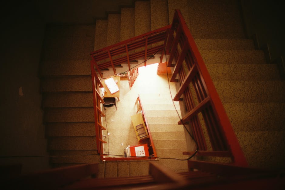 A top-down view of a narrow, indoor staircase within a Northolt home, showing beige carpeted steps bordered by a red metal handrail and balustrade. The staircase spirals downward, revealing a small landing at the bottom with various moving-related items including cardboard boxes, a wooden chair, and packaging materials. The landing area is illuminated by natural light coming from an open door or window at the bottom, suggesting this is part of a home relocation process. The surrounding walls are painted in a neutral tone, with some visible marks and scuffs. The environment indicates an interior space being prepared for furniture transport or packing and moving activities, with additional items like plastic wrapping and small packing accessories visible on the landing. This scene illustrates a typical house removal setup, captured by Man with Van Northolt during a furniture moving operation through narrow stairwells in residential settings.