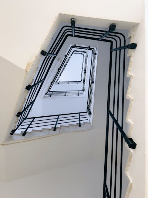A view from the bottom of a multi-story staircase looking upwards through an open rectangular shaft, with a black metal railing running along each level's edge. The stairs are made of light-colored material, possibly wood or concrete, with some steps visible along the sides of the opening. The space is enclosed by white walls, and the staircase is illuminated by natural light from above. This vertical shot captures the architectural design of the staircase, which is part of a residential property undergoing home relocation, with the focus on the structural elements and the safety railing for moving or packing processes. Man with Van Northolt may use such detailed visuals to demonstrate their expertise in navigating challenging communal staircases during furniture transport or house removals.