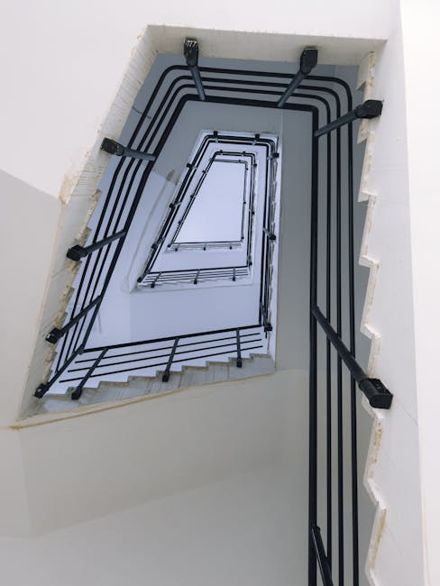 A top-down view of a narrow, indoor staircase within a Northolt home, showing beige carpeted steps bordered by a red metal handrail and balustrade. The staircase spirals downward, revealing a small landing at the bottom with various moving-related items including cardboard boxes, a wooden chair, and packaging materials. The landing area is illuminated by natural light coming from an open door or window at the bottom, suggesting this is part of a home relocation process. The surrounding walls are painted in a neutral tone, with some visible marks and scuffs. The environment indicates an interior space being prepared for furniture transport or packing and moving activities, with additional items like plastic wrapping and small packing accessories visible on the landing. This scene illustrates a typical house removal setup, captured by Man with Van Northolt during a furniture moving operation through narrow stairwells in residential settings.
