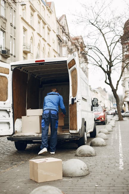 A man wearing a blue jacket and dark trousers is seen loading cardboard boxes into the back of a white removal van parked on a city street in Northolt, with residential buildings in the background and