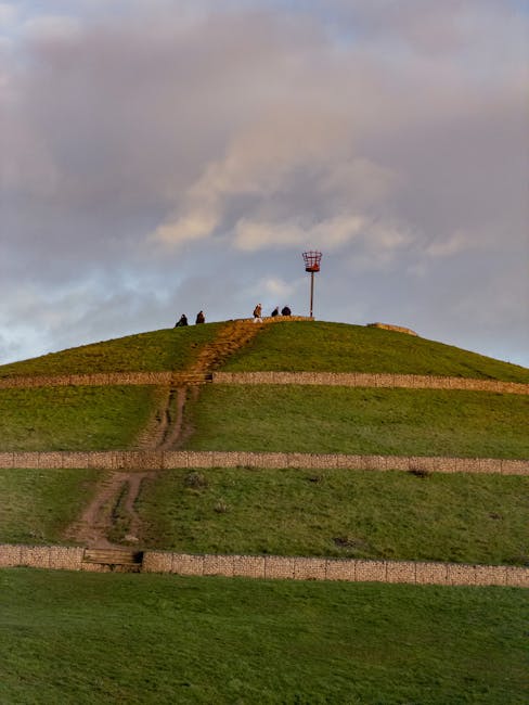A grassy, tiered hill with a worn dirt path running vertically from the bottom to the top, where several people are gathered near a red and white communication tower. The hill is divided into multiple levels by stone retaining walls, and the sky above features a mix of light and dark clouds with soft, diffused lighting. The scene depicts outdoor recreation or sightseeing in a park setting, with no interior or moving equipment visible, emphasizing the natural landscape. Natural light highlights the green grass and the textures of the pathways, supporting themes related to outdoor activities, walking, or exploring areas suitable for house removals or relocation logistics, as discussed on the page about parking and access tips for Northala Fields.