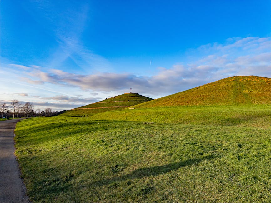 View of the grassy hills at Northala Fields in Northolt under a clear blue sky with scattered clouds. The foreground features a wide, well-maintained lawn with lush green grass, with a paved pathway visible on the left side curving into the distance. The three mounds, resembling ancient burial mounds, are covered with grass and rise gradually from left to right, with the highest hill at the top center, which has a small structure or antenna at the summit. The bright sunlight casts gentle shadows on the slopes, highlighting the contours of the mounds. This scenic landscape is often used as a background for local community events and outdoor activities, and is relevant to recent house removal or furniture transport activities carried out by local removals companies such as Man with Van Northolt, especially when accessing the site for parking and loading purposes during home relocations.
