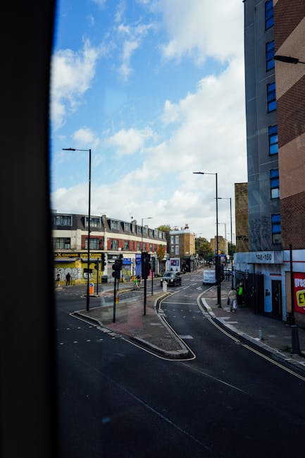 View of a busy urban street scene from inside a vehicle, showing a curved road with marked lanes and a pedestrian crossing. On the left, a row of commercial buildings includes a currency exchange shop with a yellow sign and exterior advertising. Several pedestrians are walking along the sidewalks or waiting at the crossing, and a few vehicles are visible on the road, including white vans and cars. Streetlights and overhead wires are present, and the sky above is partly cloudy with patches of blue. The scene depicts a typical Northolt area, relevant to moving services by Man with Van Northolt, as part of home relocation or furniture transport preparations. Adjacent to the sidewalk, a building with a brick facade and large windows can be seen on the right, and additional buildings further down the street contribute to the urban environment where local removals are commonly carried out.