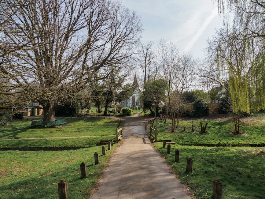A paved pathway in a park or garden setting, bordered by short wooden posts on each side, leading towards a small wooden footbridge with railings. The pathway continues beyond the bridge, surrounded by well-maintained grass, trees, and shrubs. Some trees are leafless, indicating early spring or late autumn, while others have green foliage or hanging branches. In the background, a historic church with a pointed steeple is visible among the trees, under a partly cloudy sky with some high thin clouds. The scene is illuminated by natural daylight, creating shadows on the ground. This tranquil outdoor environment may be used as part of a house removal process involving careful packing and transportation of household items stored in the area, with [COMPANY_NAME] ensuring professional moving logistics and furniture transport services.