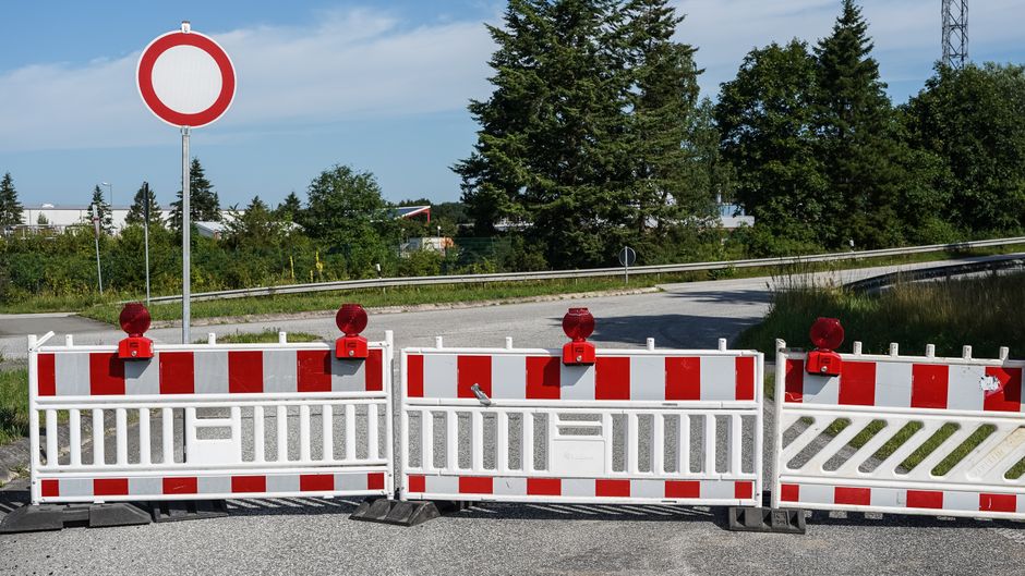 Photograph of a road closure with red and white striped barriers and red warning lights, set up in front of an intersection during daytime. Behind the barriers, a van is partially visible, parked on the pavement near a residential area with green trees and bushes. The background shows a clear sky with some scattered clouds and a metal guardrail along the road. This scene reflects temporary parking restrictions and traffic management measures that may be encountered during house removal or furniture transport operations, emphasizing the importance of planning and adherence to road closure rules for efficient home relocation. Such barriers and signage are part of the logistics managed by a professional removals service like Man with Van Northolt, ensuring safe loading, unloading, and transport of household items amidst road restrictions.
