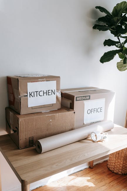 Inside a residential room, three cardboard boxes labeled 'Kitchen' and 'Office' are stacked on a light wooden table, indicating packing and moving preparations. The boxes are sealed with clear packing tape, with one box marked 'Medium' and another displaying the word 'Kitchen' in bold black letters. A white roll of packing paper or bubble wrap is positioned on the table, alongside a small, partially unrolled sheet of paper or tape. To the right, a small roll of packing tape and a pair of scissors are visible, used in the packing process. The scene is illuminated by natural light from an unseen window, with a large green houseplant partially visible in the background, adding a touch of greenery to the setting. The environment suggests a home undergoing a move or packing for a relocation, which aligns with the services offered by Man with Van Northolt, specialising in removals and home relocation logistics, particularly for urgent tenancy move-outs in Northolt.
